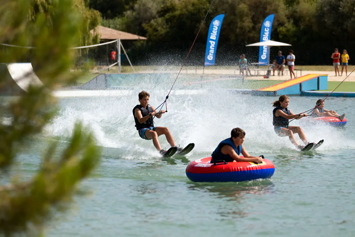 Personnes pratiquant le ski nautique et la bouée tractée sur un lac, avec public et bannières en arrière-plan.