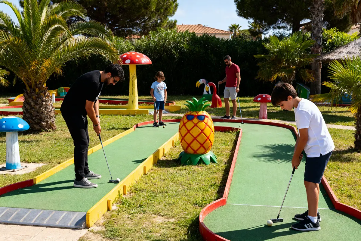 Personnes jouant au mini-golf dans un parc, décorations colorées de fruits et champignons autour.