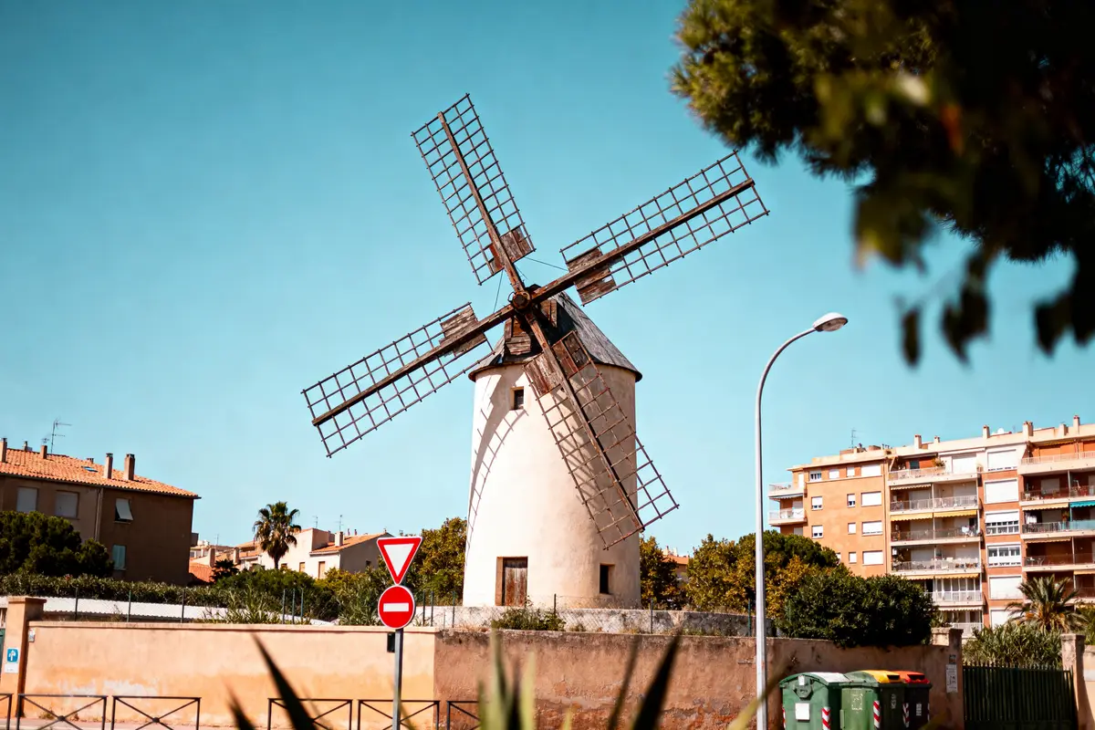 Moulin traditionnel entouré de bâtiments résidentiels, ciel bleu, panneaux de signalisation, et arbres verdoyants.